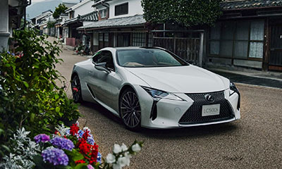 A white Lexus LC driving down a flower-lined road.