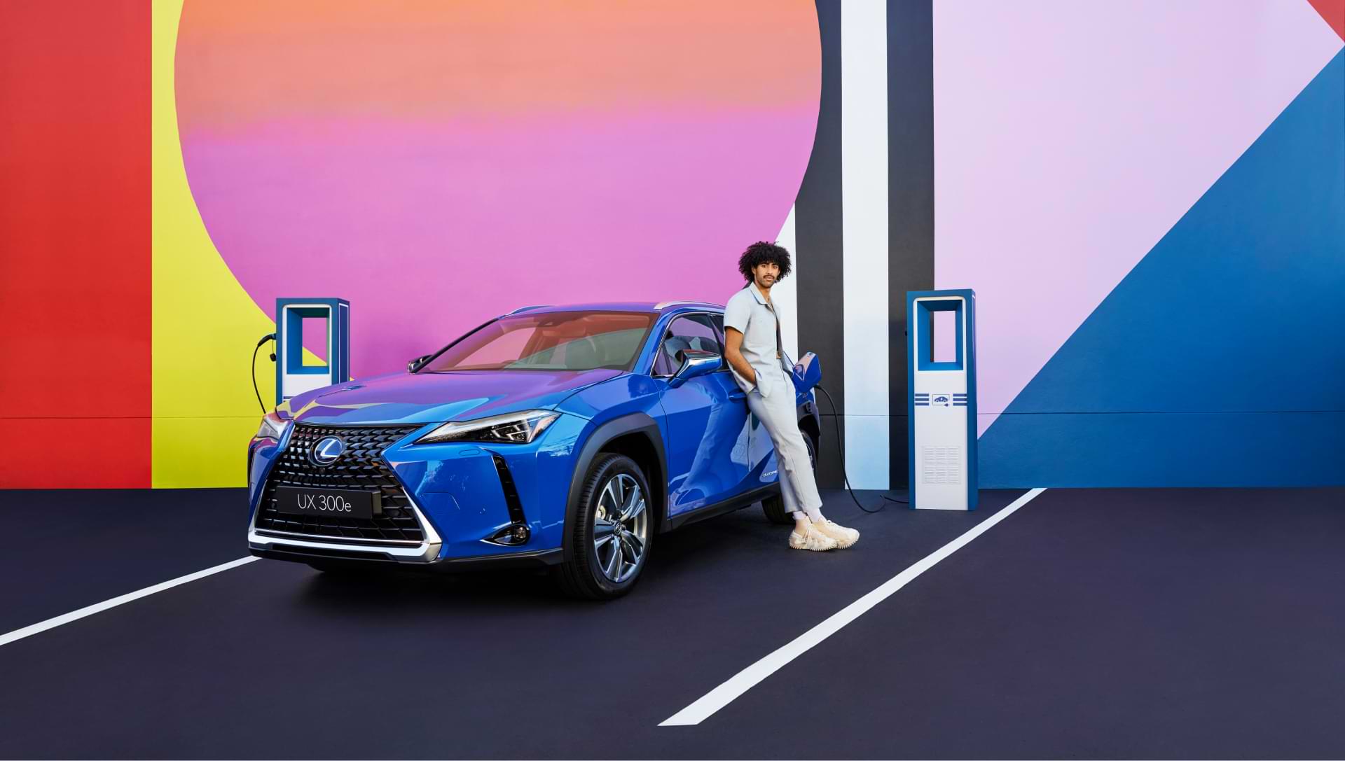 A man in a light blue suit leans against a bright blue UX 300e as it charges at a station. The background wall is brightly coloured pinks and yellows.