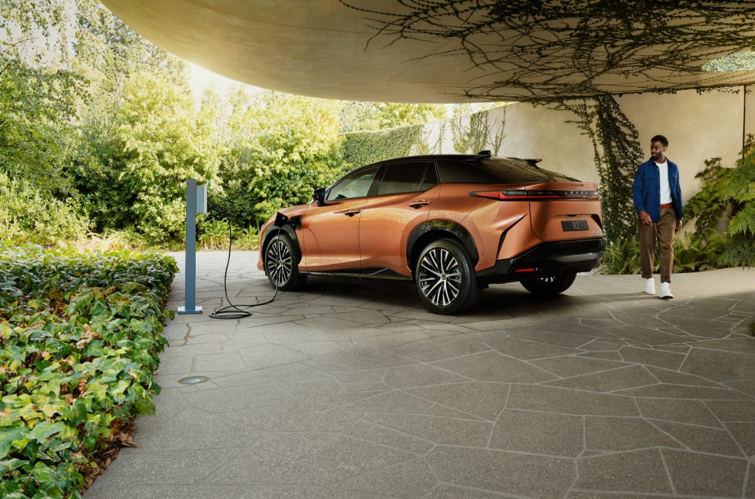 An orange Lexus RZ is parked and charging in a leafy green, vine-filled driveway. A man is seen walking around the rear of the car.