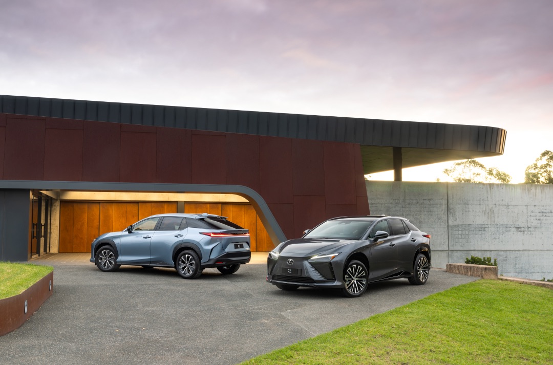A blue and dark grey Lexus RZ are parked in the concrete driveway of a large concrete structure. Their lights are on.