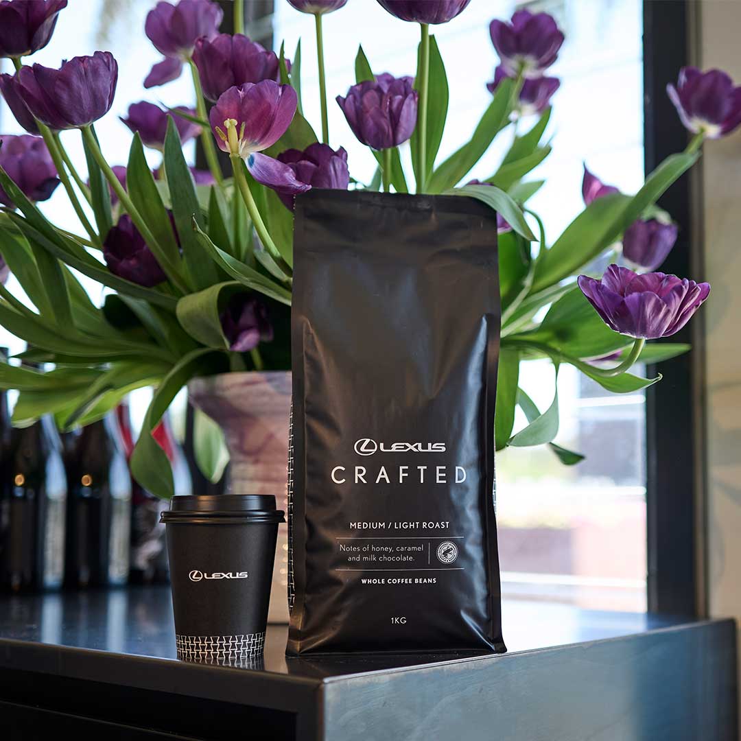 A bag of St Remio x Lexus branded coffee beans on black topped table. A cup of coffee and a white vase holding purple flowers sit alongside.