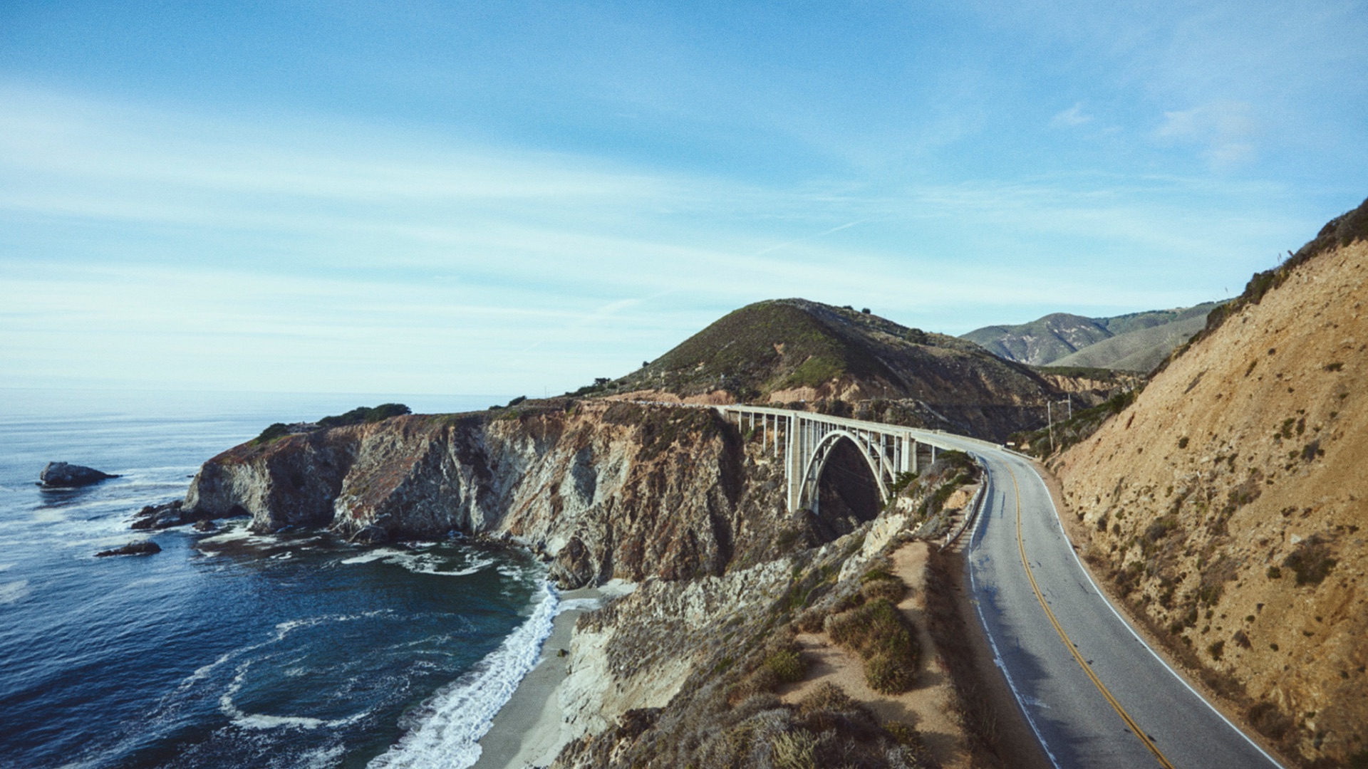 An aerial view of a road winding around a cliff, waves crashing to the left, cliffs to the right.