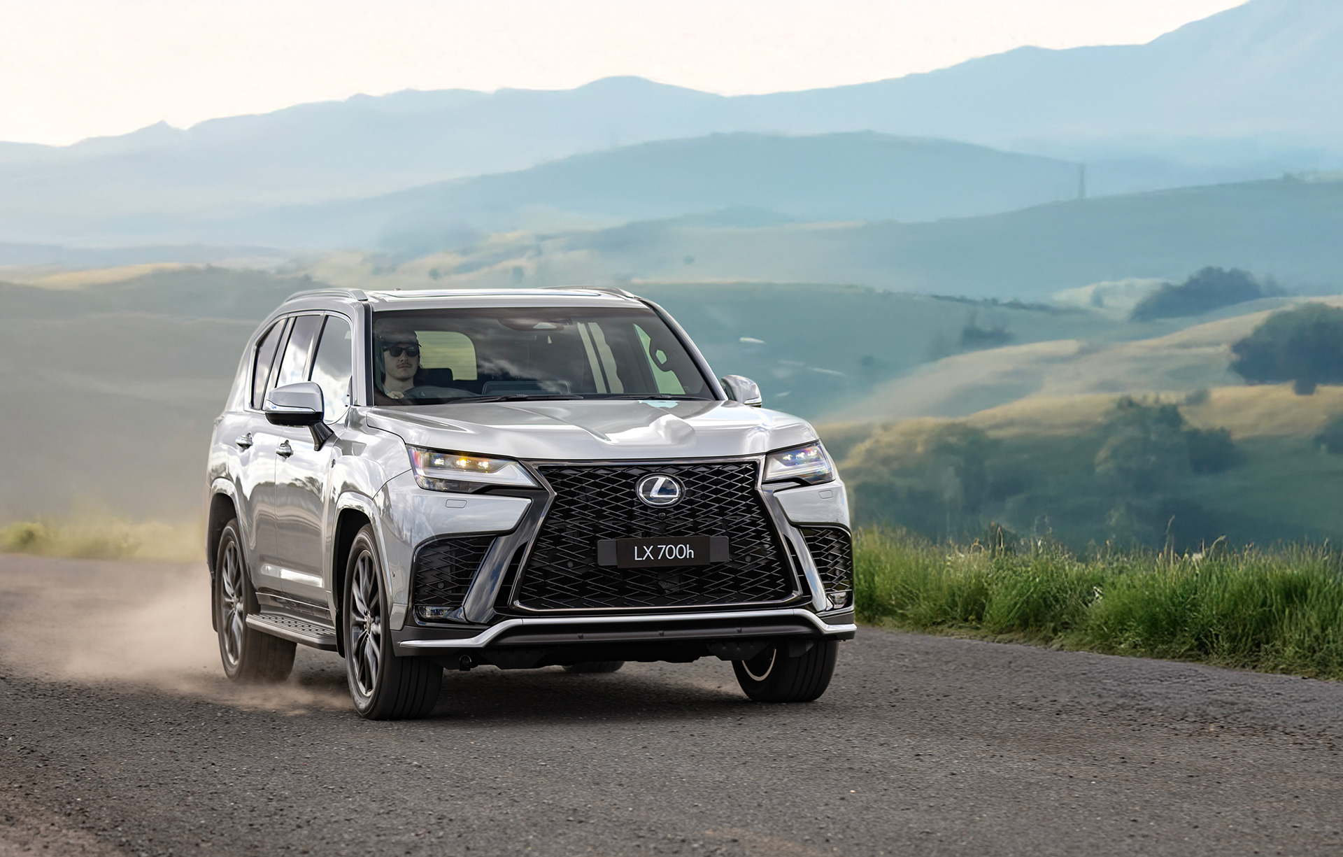 Lexus LX 700h driving along a winding country road with mountains in the background.