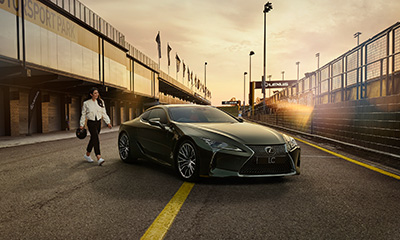 A woman walks towards a Lexus LC coupe parked in a motoring pit lane.